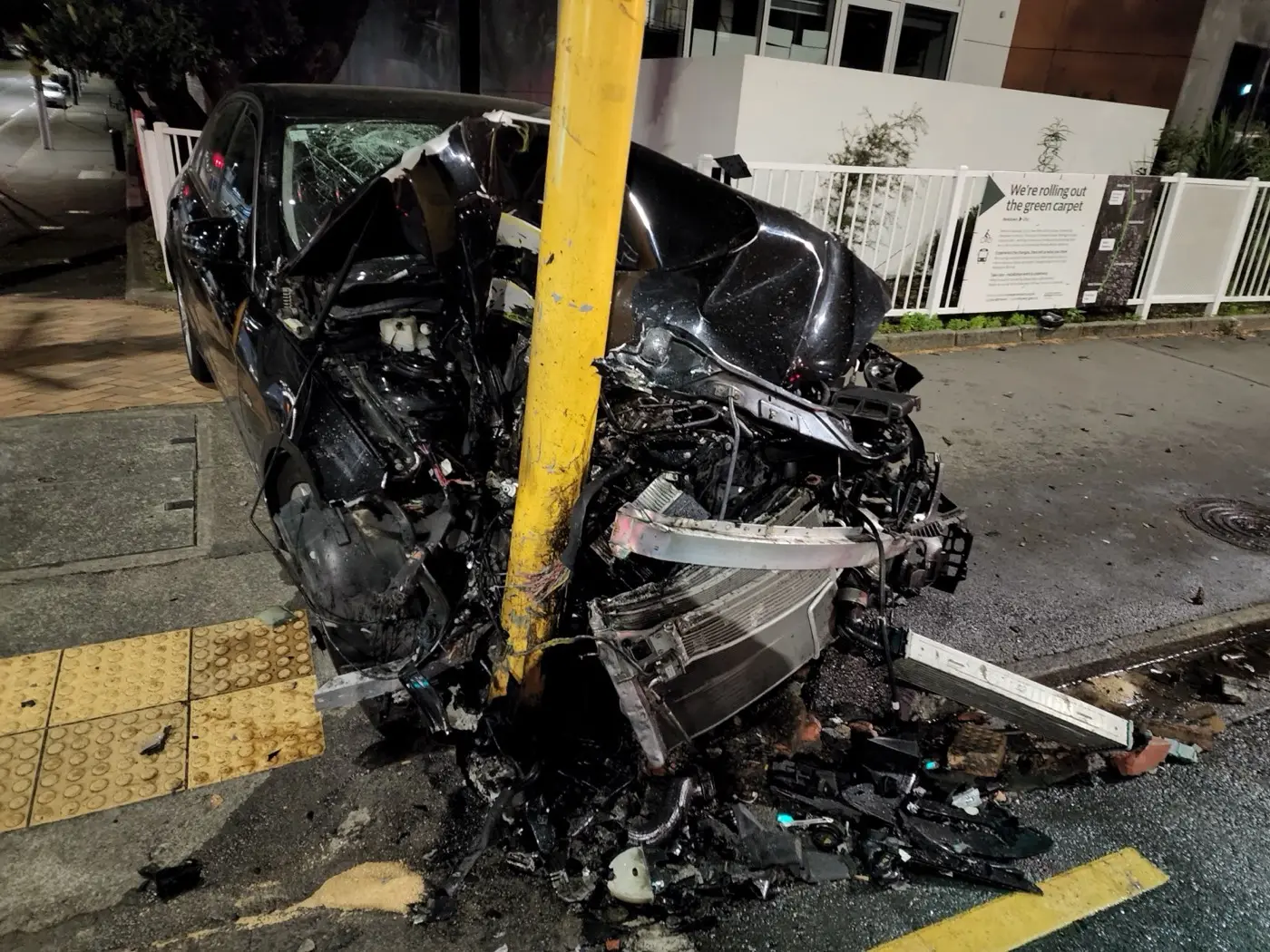 A black car heavily damaged at the front from an impact with a pole at a pedestrian crossing.