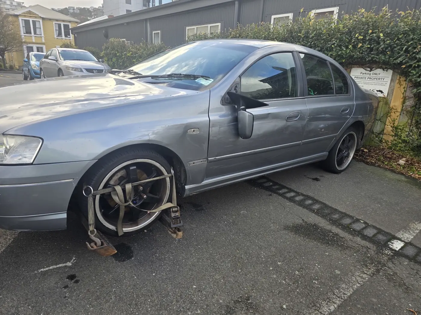 A grey car with its front wheels lifted by a tow truck, in front of a sign indicating a private parking area.