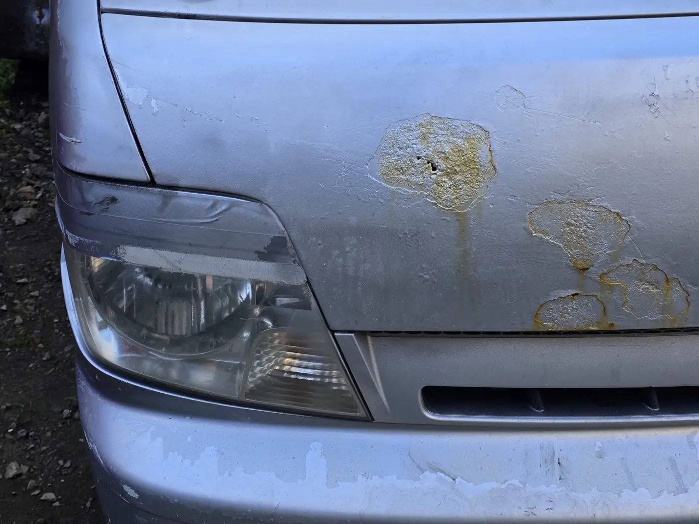 The front-right corner of a silver vehicle, with visible rust and holes through the steel panel.